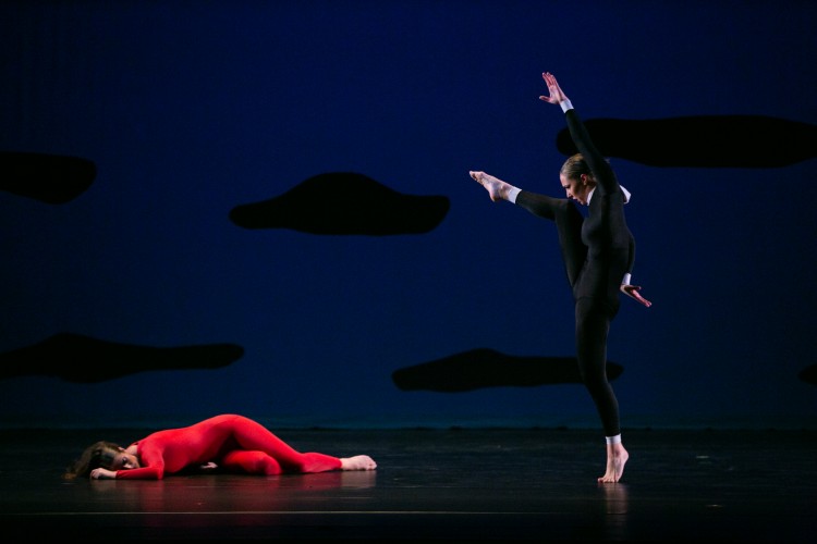a woman in a red unitard lies on the floor as another woman dressed in a black unitard with a white collar and white cuffs hovers over her menacingly